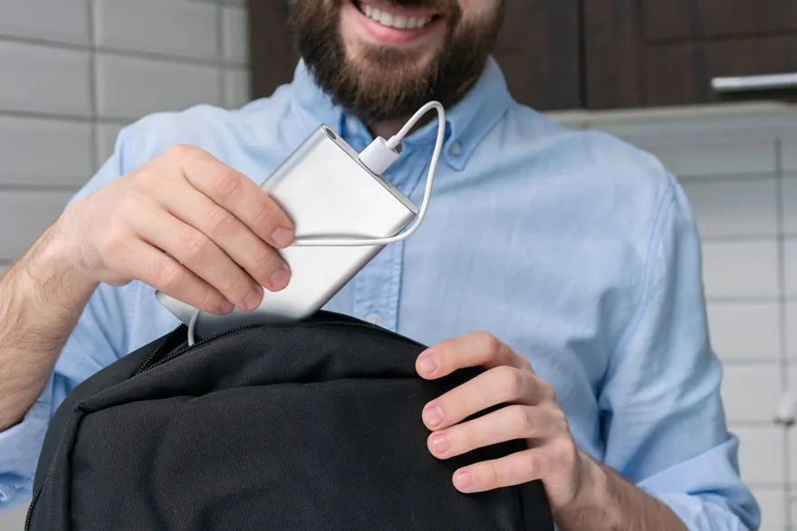 Un chargeur argenté est mis dans un sac à dos noir. Un cordon blanc le relie. L'arrière-plan est carrelé de blanc.