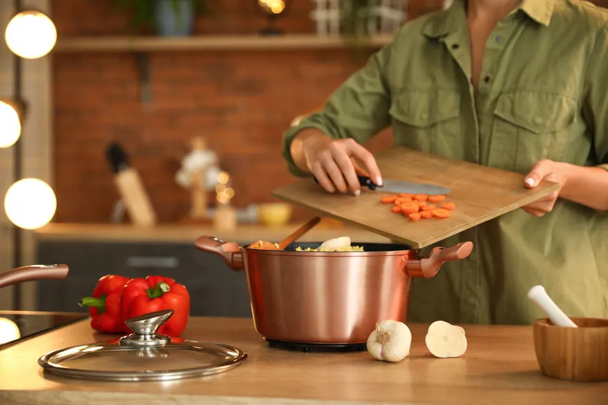Een keukenopstelling met een koperen pot, rode paprika's, knoflook en een snijplank met wortelen.
