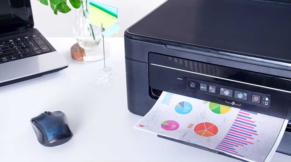 A black printer is printing a page with colorful charts. A laptop and mouse sit on a white desk, with a plant in a vase behind.