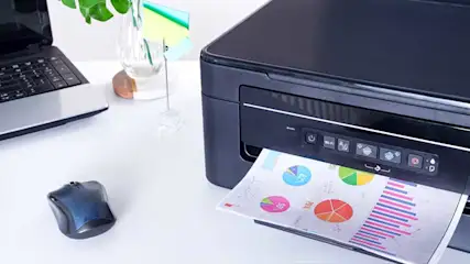 A black printer is printing a page with colorful charts. A laptop and mouse sit on a white desk, with a plant in a vase behind.