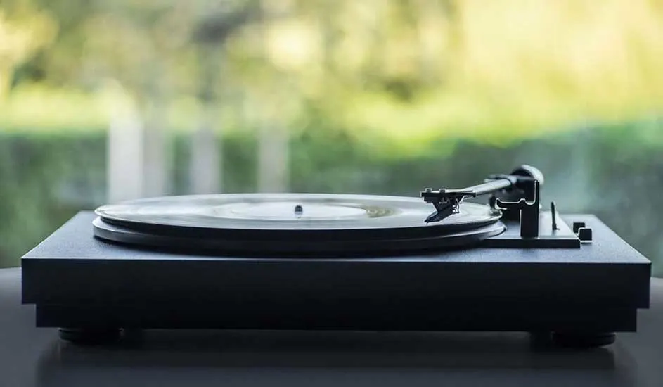 Platine noire avec disque vinyle sur une surface grise, devant un arrière-plan flou de verdure.