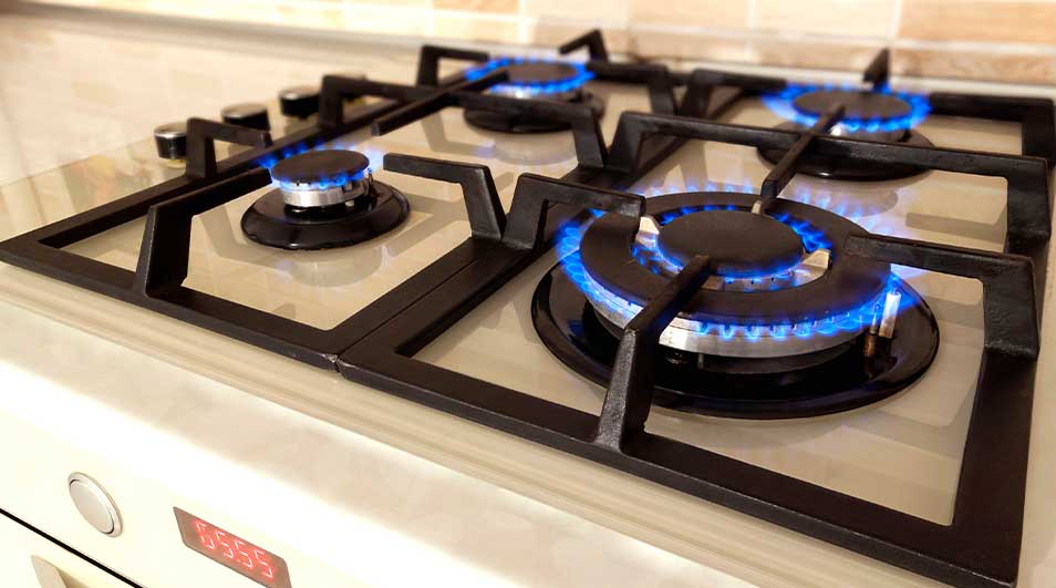 Een close-up van een gasfornuis met vier branders aan. Blauwe vlammen branden onder zwarte pannenroosters op een licht aanrecht.