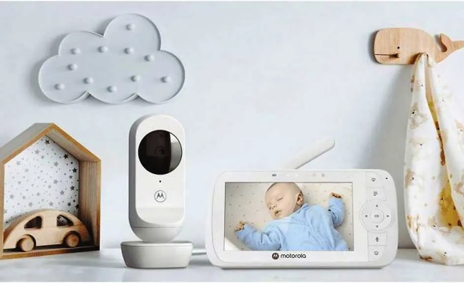 Een babyfoon met camera en monitor staat op een witte plank met decoratieve objecten tegen een witte muur.