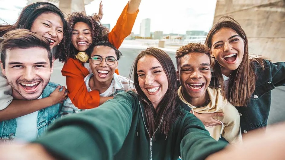 Un groupe de personnes souriantes prend un selfie devant un mur gris et des bâtiments urbains. Les couleurs vives des vêtements ressortent.