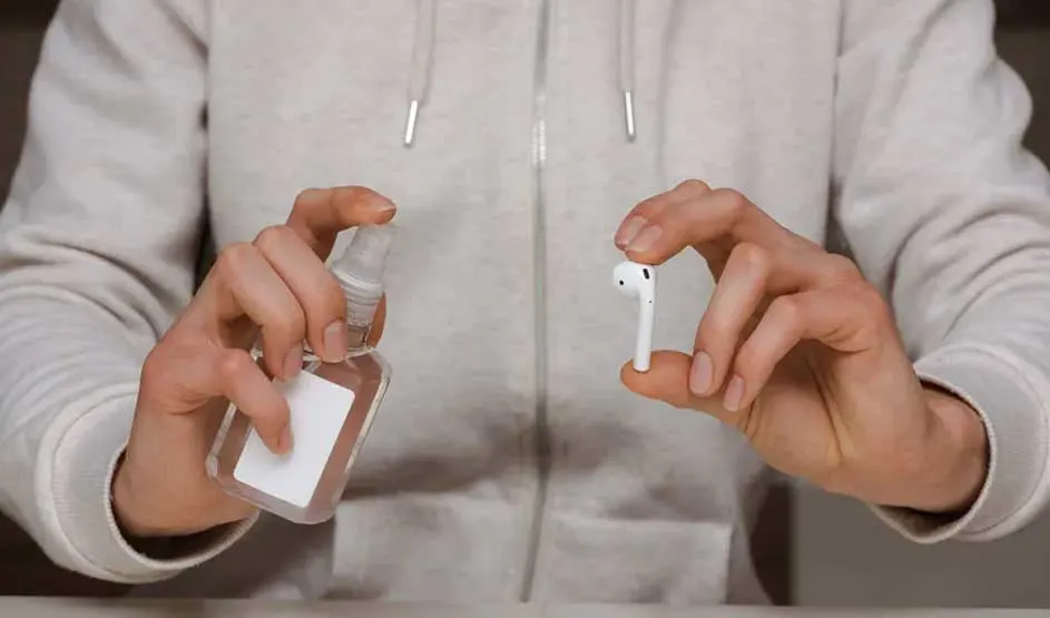 Een persoon houdt een witte draadloze oortelefoon en een doorzichtige sprayflacon vast tegen een grijze hoodie-achtergrond.