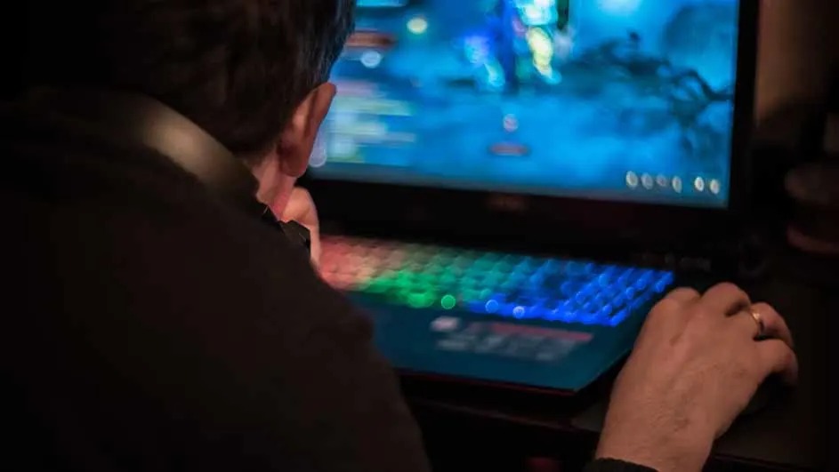 Een laptop met een verlicht toetsenbord toont een blauw videospel op het scherm. Handen bedienen de muis.