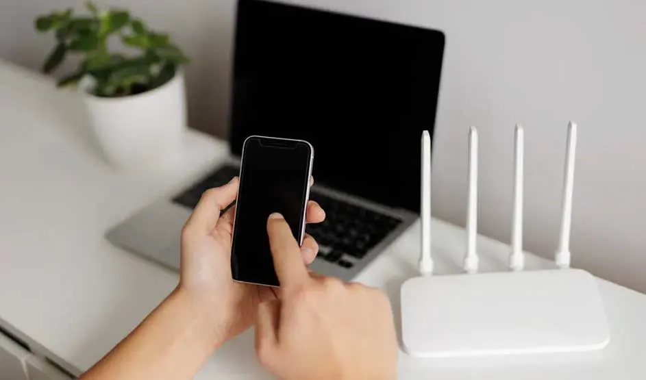 Un ordinateur portable, un routeur blanc à quatre antennes et un smartphone sur une table blanche, avec une plante en arrière-plan.