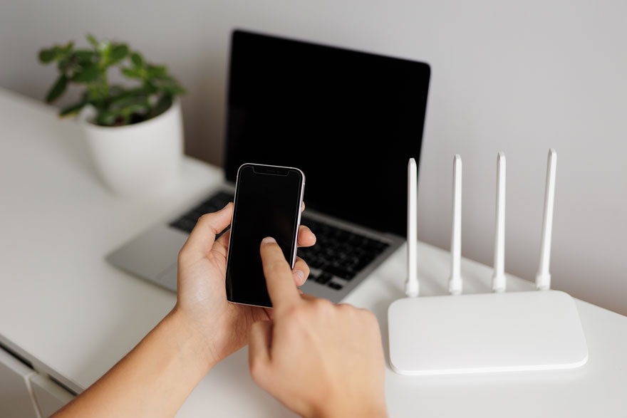 Op een witte tafel staan een laptop met een zwart scherm, een witte router met vier antennes en een plant. Een hand houdt een zwarte telefoon vast.