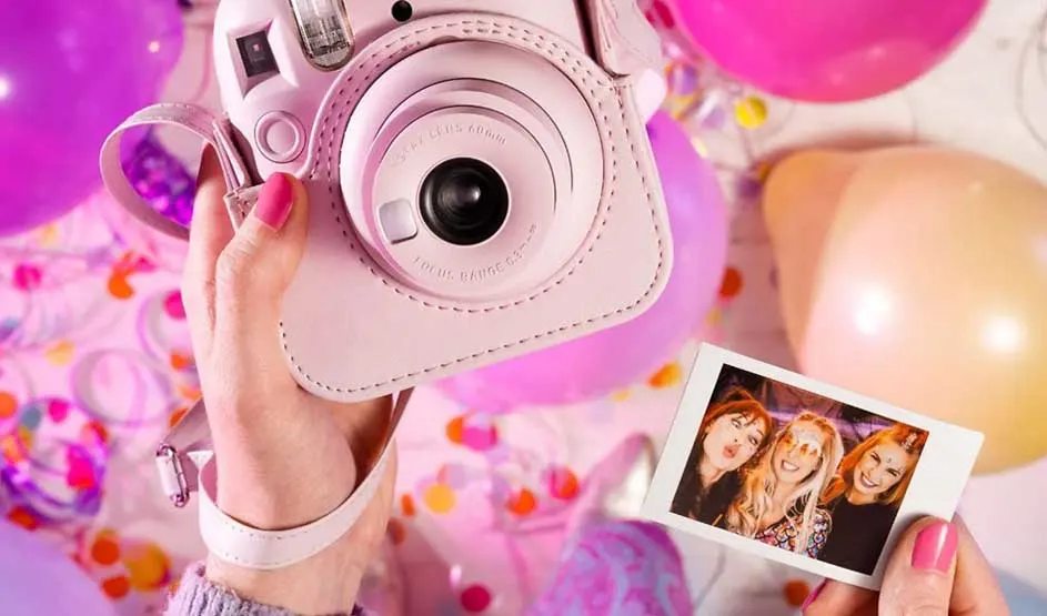 A hand holds a pink instant camera displaying a polaroid photo of women. Colorful balloons and confetti surround the festive, joyful scene in vibrant pink tones.