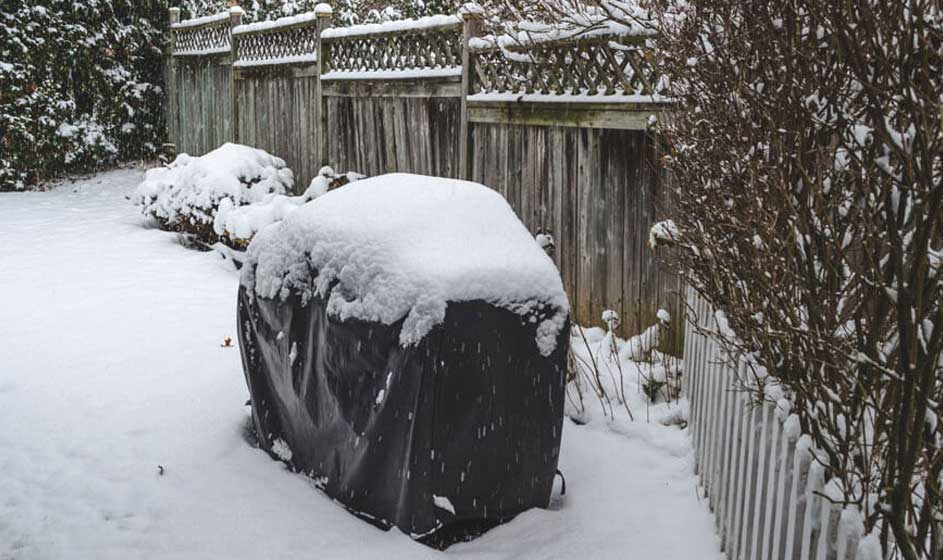 Een winterlandschap met een zwarte hoes bedekt met sneeuw op de voorgrond. Achtergrond: houten schutting en besneeuwde struiken.