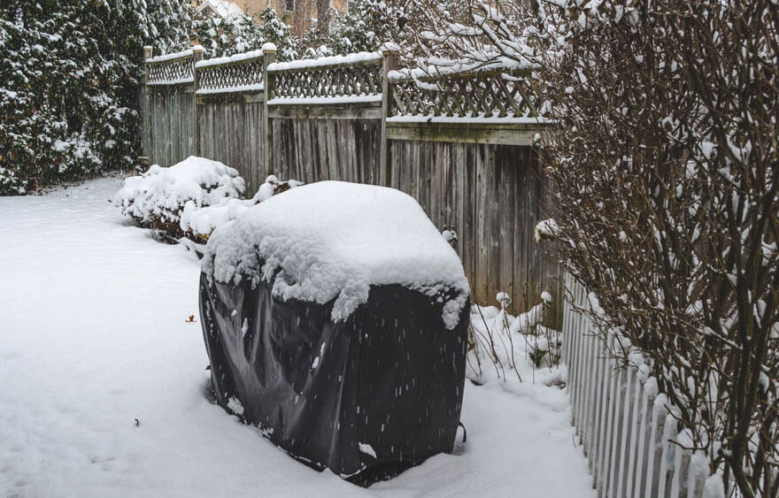 Un jardin enneigé avec une housse noire sur un objet, des buissons et des clôtures en bois sous la neige.