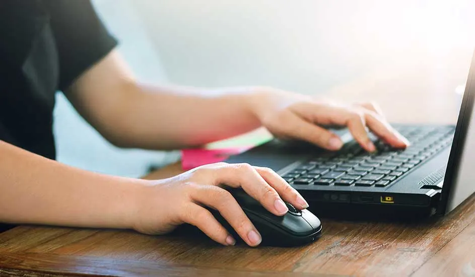 Een zwarte laptop en muis op een houten tafel, met handen die typen. Heldere achtergrond.