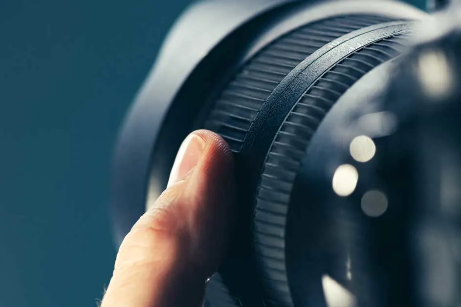 Een vinger draait aan de geribbelde ring van een zwarte cameralens tegen een blauwe achtergrond. De lens reflecteert lichtplekken.
