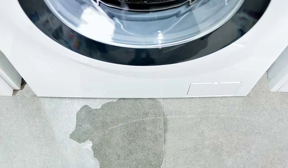 Een close-up van een witte wasmachine op een grijze tegelvloer met een plas water ervoor.
