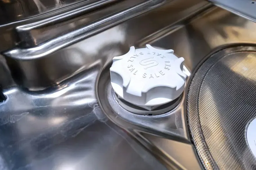 Een close-up van het zoutreservoir en het metalen filter in een vaatwasser. Er is geen merk zichtbaar op het apparaat.