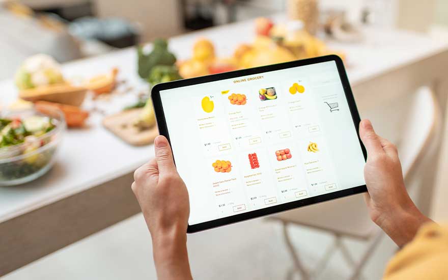 Someone is holding a tablet with an online groceries shop displayed, sitting at a kitchen table with fruits in the background.