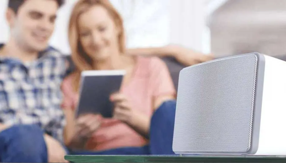 Un haut-parleur blanc sur une table verte, avec un écran gris en maille. À l'arrière-plan, une femme tient une tablette.