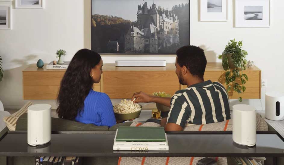 Twee mensen zitten op de bank met een kom popcorn, kijkend naar tv, omringd door witte Sonos speakers in een modern ingerichte woonkamer.