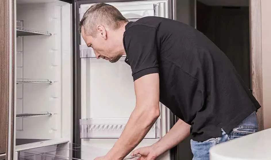Een open, witte koelkast met zwarte rand staat in een kamer met een houten kast en deur. De koelkast heeft meerdere planken en een transparante lade.