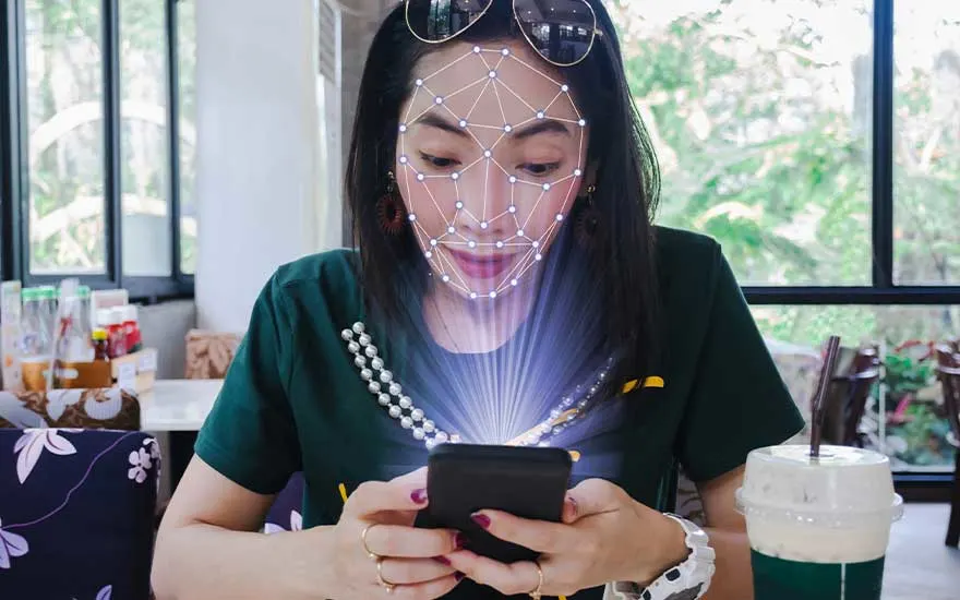 Une personne utilise un smartphone avec un quadrillage facial numérique en superposition. Une boisson et une fenêtre sont visibles.