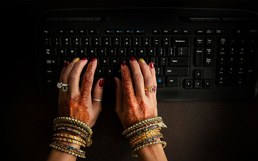 Twee handen met henna-tatoeages en veel armbanden typen op een zwart toetsenbord tegen een donkere achtergrond.