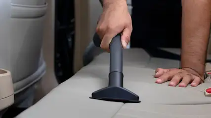 Een hand houdt een grijze stofzuiger vast op een beige autostoel. De auto heeft beige interieur.