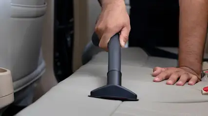Un aspirateur gris nettoie un siège de voiture beige. La main guide l'appareil. L'arrière-plan est flou, montrant l'intérieur d'une voiture.