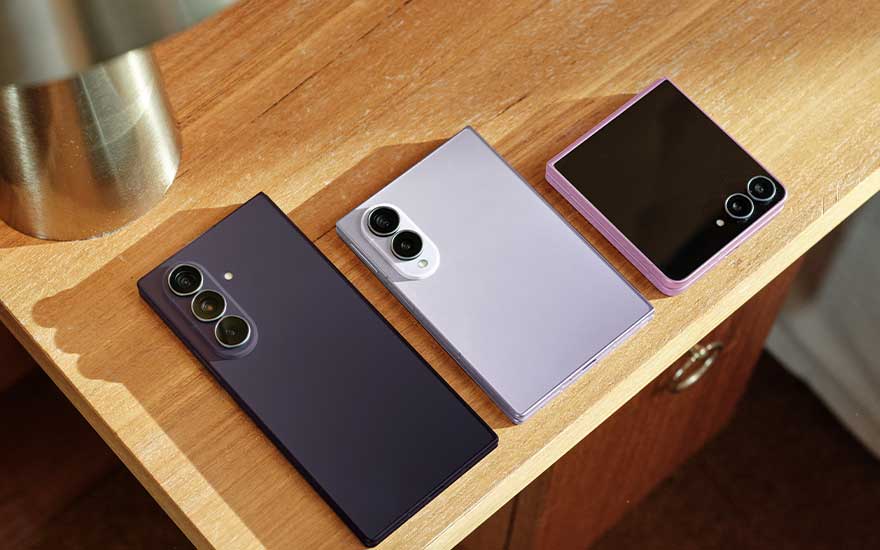 Trois smartphones pliables Samsung sont posés ouverts et fermés sur une table en bois, près d’une lampe métallique.