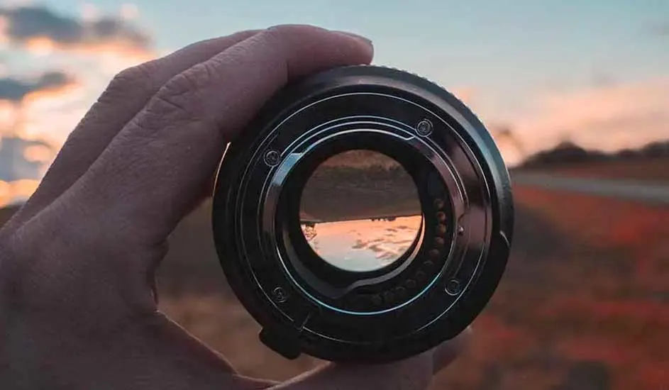 Een hand houdt een cameralens vast. Door de lens is een zonsondergang met een oranje lucht en een bruin landschap te zien.