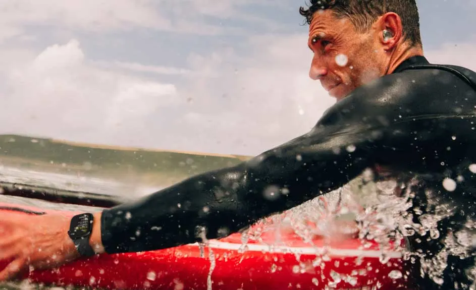 Un surfeur en combinaison noire sur une planche rouge, avec une montre à son poignet et des écouteurs, sous un ciel nuageux.