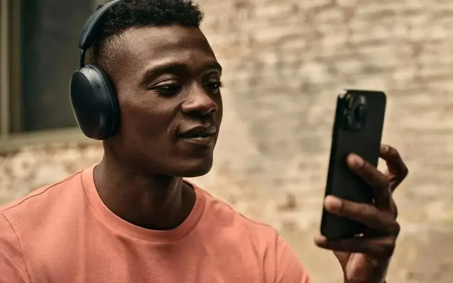 Un homme porte un casque noir et tient un téléphone noir devant un mur texturé beige.