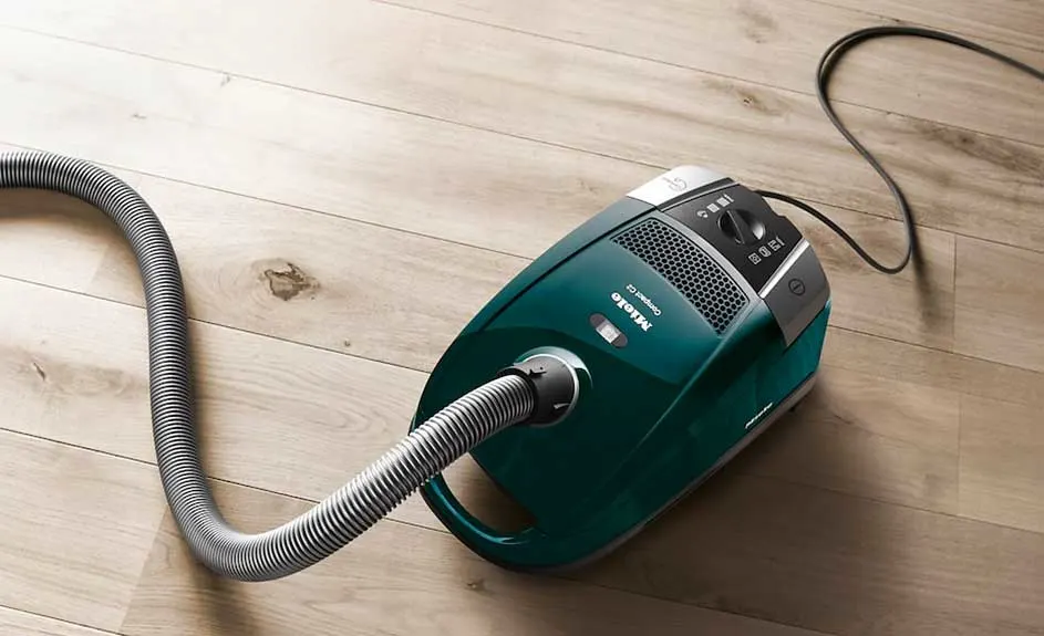 Een groene Miele stofzuiger staat op een houten vloer met een slang en een zwart snoer.