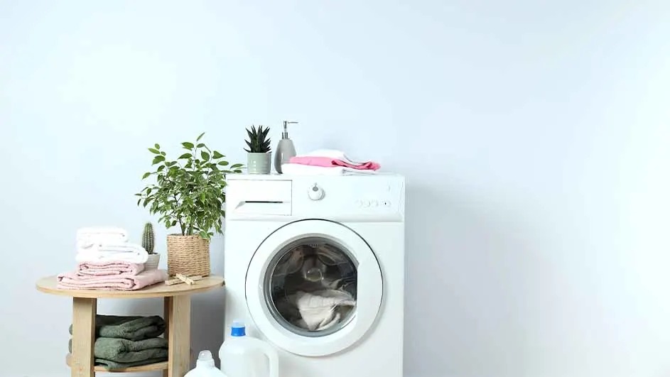 Een witte wasmachine met wasgoed in de trommel staat naast een houten tafeltje met opgestapelde handdoeken en planten, merk is niet zichtbaar.