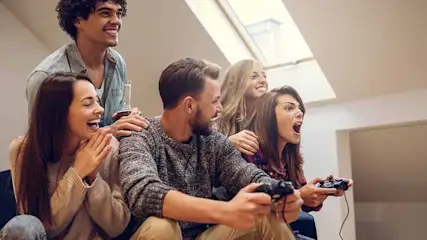 Zes mensen zitten in een kamer. Twee spelen een videospel met zwarte controllers. Er is een raam op de achtergrond.