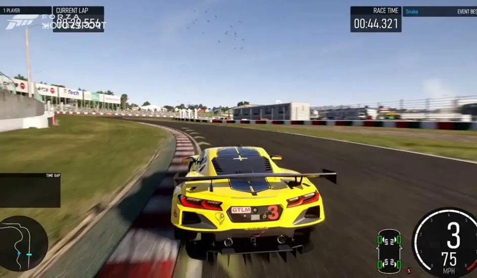 Gele raceauto op een circuit met groene bermen en gebouwen op de achtergrond, onder een blauwe hemel.