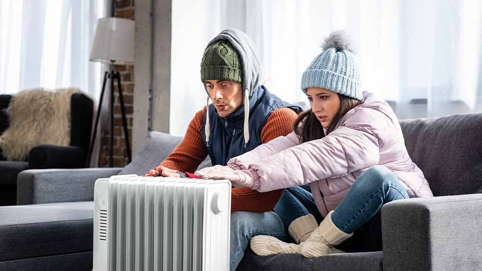 Un radiateur blanc est placé devant un canapé gris. Des vêtements chauds sont portés, comme des bonnets et des vestes.