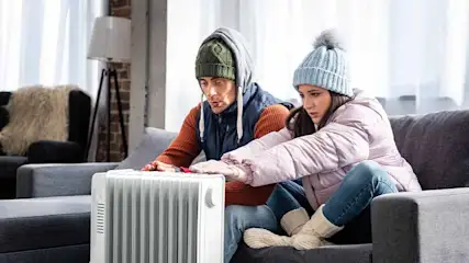 Un radiateur blanc est placé devant un canapé gris. Des vêtements chauds sont portés, comme des bonnets et des vestes.