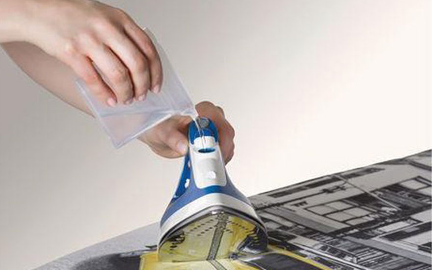 Main tenant un sac transparent, une personne remplit un fer à repasser bleu et blanc posé sur une planche à repasser à motifs.