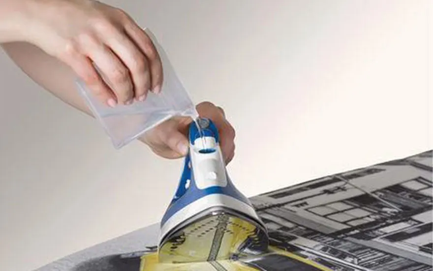 Main tenant un sac transparent, une personne remplit un fer à repasser bleu et blanc posé sur une planche à repasser à motifs.