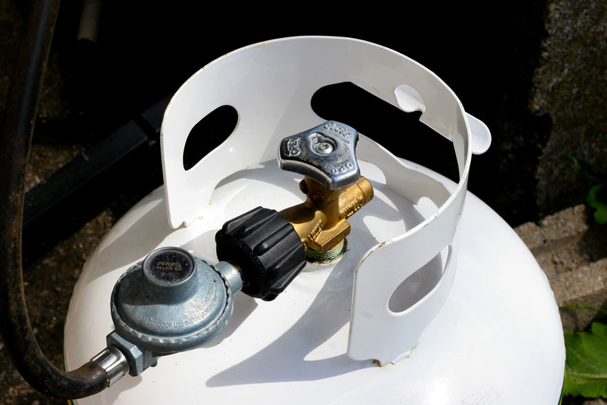 Een witte propaantank met een zwarte slang, een grijze regelaar en een gouden ventiel tegen een donkere achtergrond.