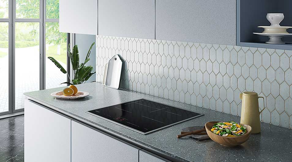 Cuisine moderne: comptoir en granit, plaques de cuisson noires, crédence à motifs, bol de salade, pichet beige. Fenêtre et plante à gauche.