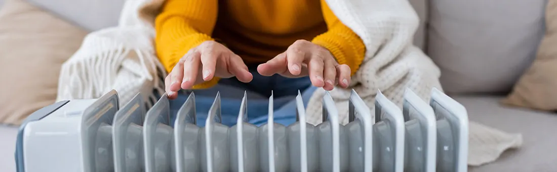 Gros plan d'un radiateur à huile blanc sur un canapé gris avec des mains tendues au-dessus.