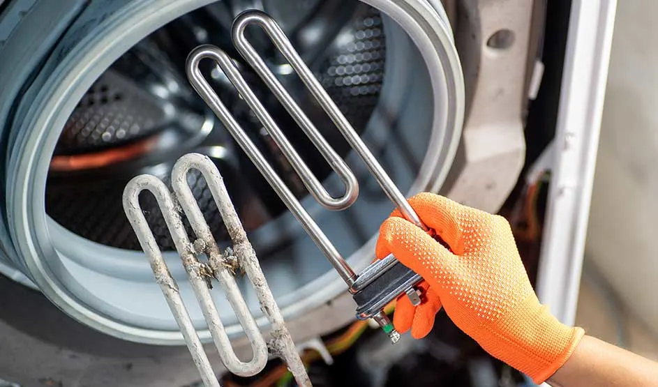 Une main en gant orange tient un élément chauffant neuf et un entartré devant un lave-linge, la marque n’est pas visible.