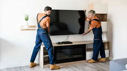 Deux hommes en salopette bleue installent un téléviseur noir sur un meuble TV beige avec une cheminée noire.