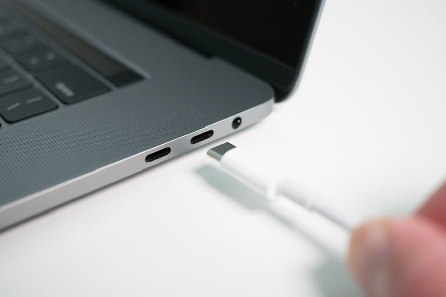 Een grijze laptop met zwarte toetsen en een witte USB-C kabel die in een van de poorten wordt gestoken op een witte ondergrond.