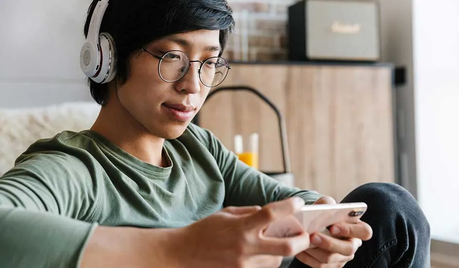 Un téléphone dans les mains, des écouteurs blancs, un t-shirt vert, un pantalon noir et un haut-parleur beige en arrière-plan.