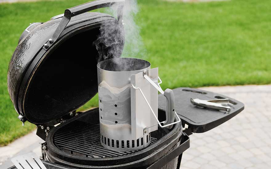 Een zwarte barbecue met open deksel, een zilveren brikettenstarter met rook en een groene grasachtergrond.