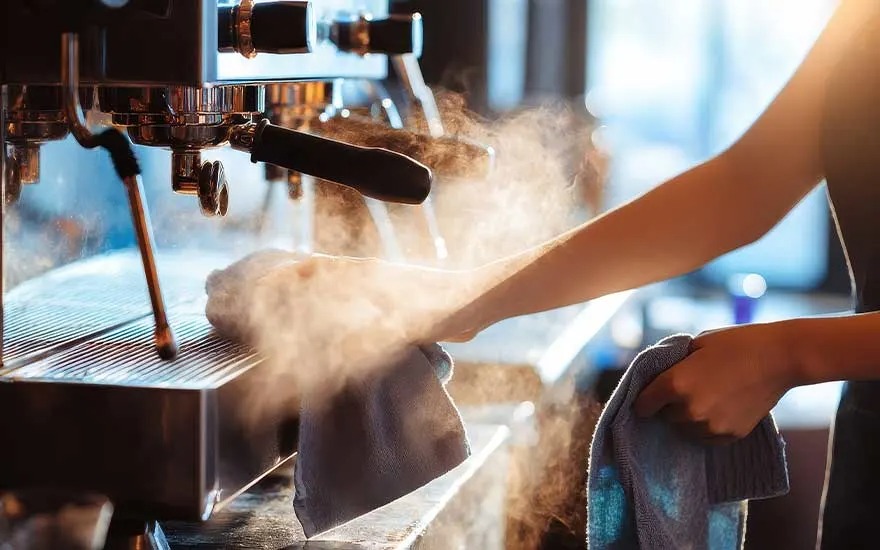 Des mains essuient une machine à espresso fumante avec un chiffon, la vapeur monte dans l’ambiance chaleureuse du bar.