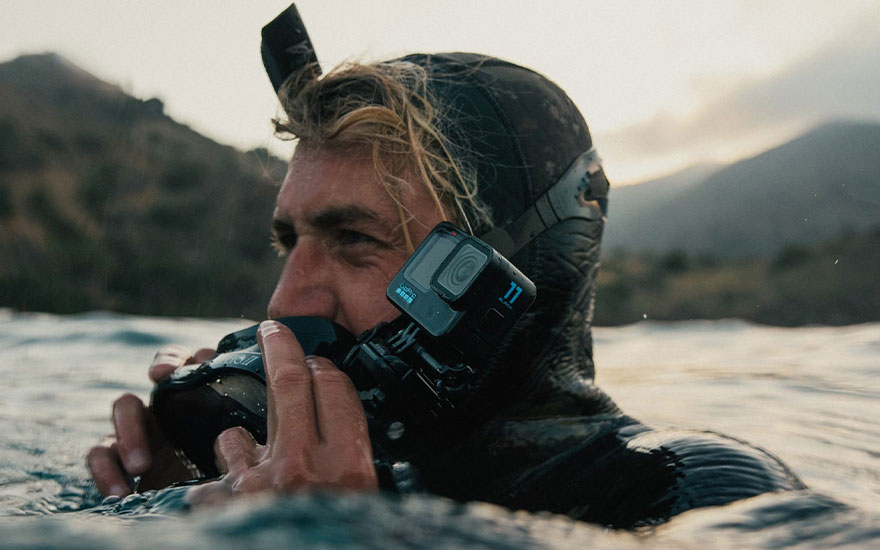 A black GoPro camera is attached to a black diving mask in water. Mountains are visible in the blurred background.