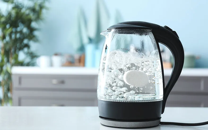 Een glazen waterkoker met zwarte accenten staat op een wit aanrecht. Binnenin kookt water. Op de achtergrond staat een grijs kabinet.